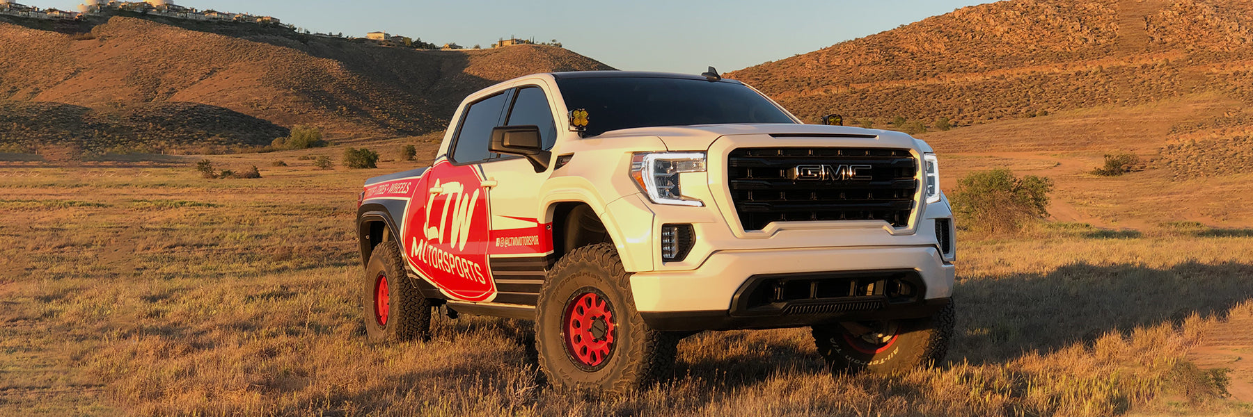 A white GMC off-road truck with red accents and decals parked on a grassy field during sunset. The rugged vehicle is equipped with large red wheels, and the landscape features gentle rolling hills under a clear sky.